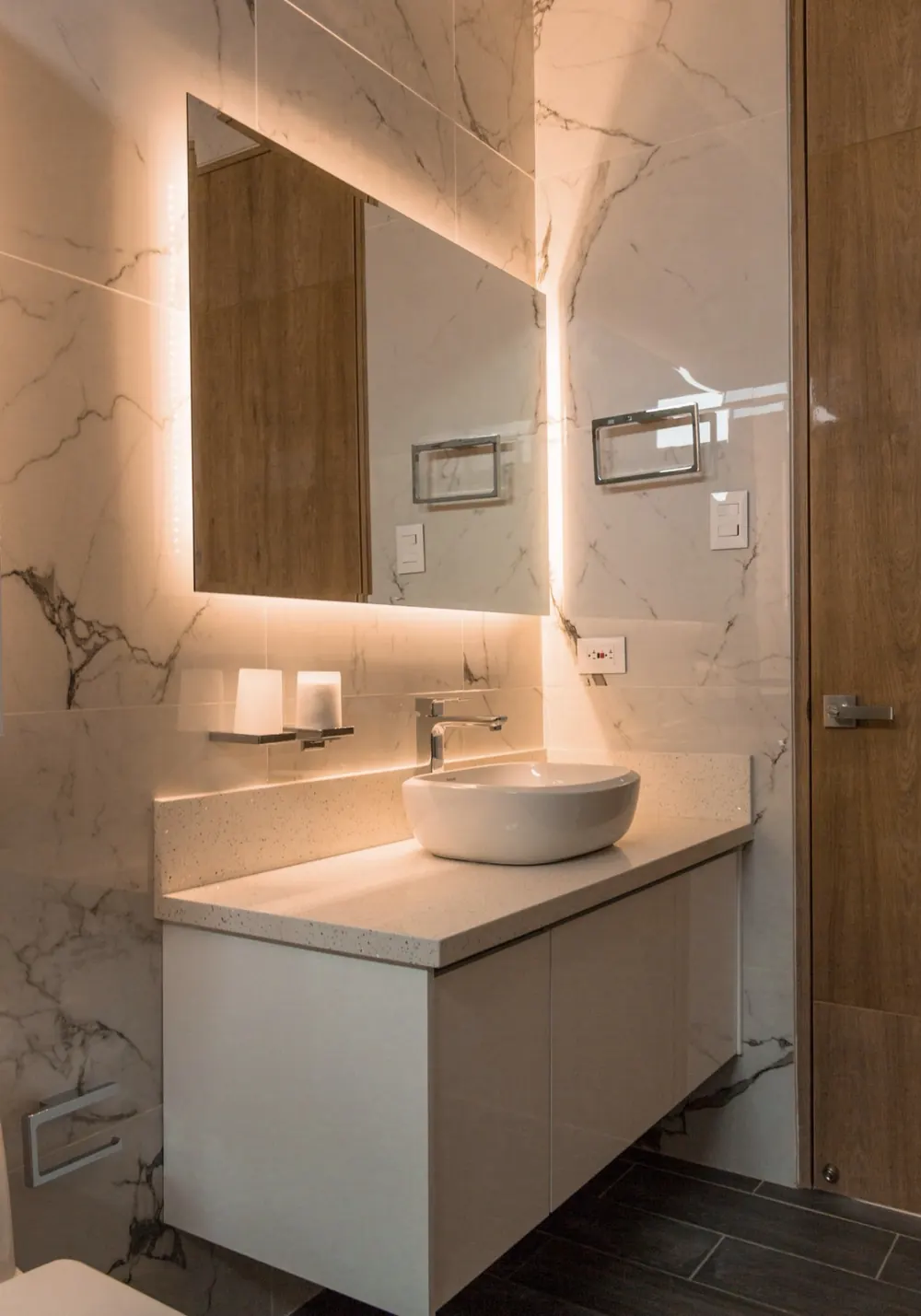 Baño con espejo con luz perimetral, lavabo sobre encimera y revestimiento efecto mármol